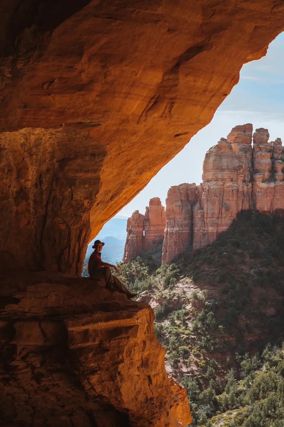 Evan in a hat sitting on a ledge inside the Keyhole Cave, red rock spires beyond