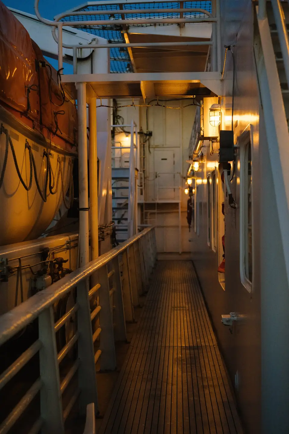 A lamplit side deck of a ship, lifeboats hanging overhead, near Juneau, Alaska