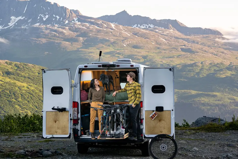 Evan, Melanny, and Penelope in the open van doors with a mountain bike, alpine ridges behind, near Valdez