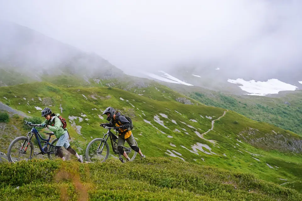 Two mountain bikers pushing their bikes up a green alpine slope under low cloud near Alyeska, Alaska