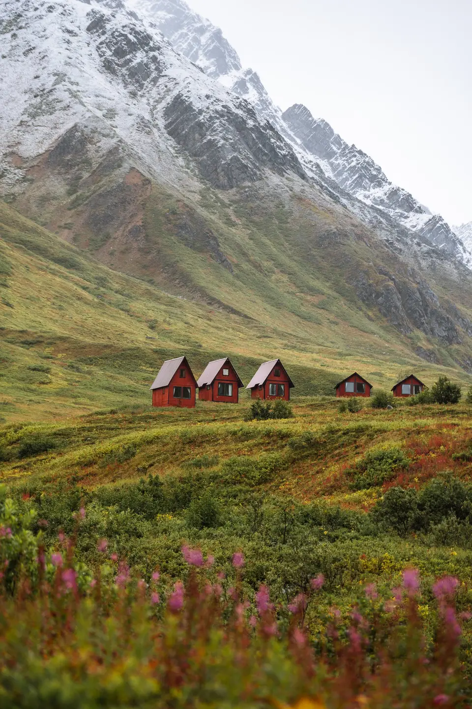 Five red cabins on a green slope below a snow-dusted peak at Hatcher Pass, Alaska
