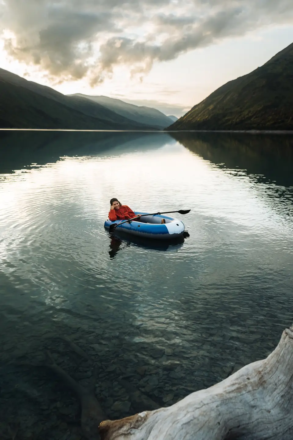 A paddler in a blue packraft on a still mountain lake at dusk on the Kenai Peninsula, Alaska