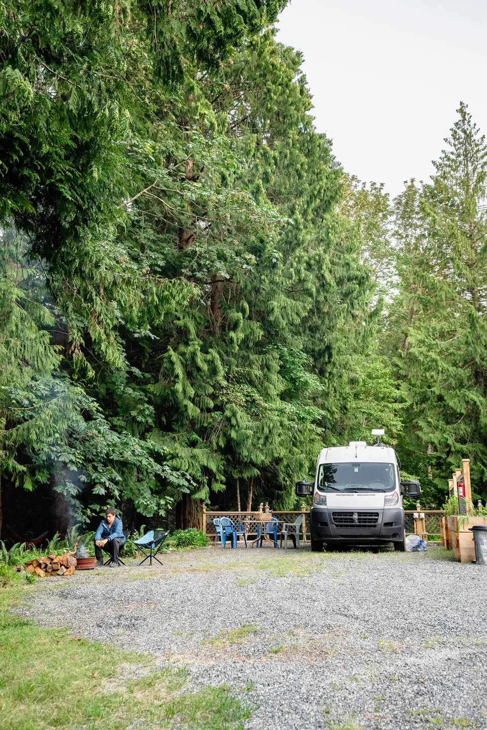 A person sitting near a campfire in a natural setting with tall pine trees, an outdoor table with chairs, and a parked white van.