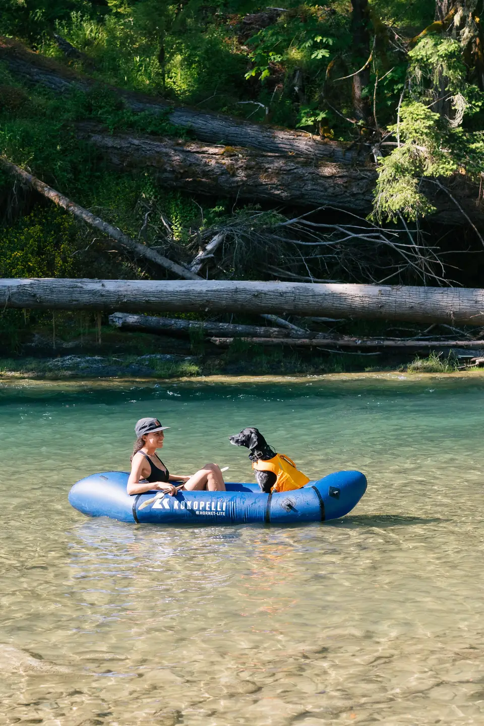 A woman and her dog in a blue inflatable boat on a clear, shallow river with a forested bank and fallen logs in the background.