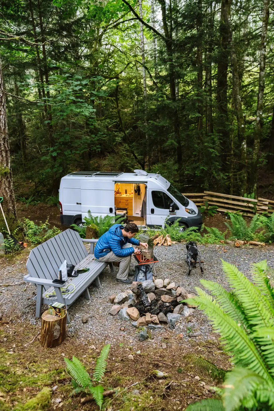 A person sitting on a gray bench in a forested campsite, tending to a small portable grill. A black and white dog is nearby. A white van with its door open is parked behind the person, and a fire pit with stones surrounds a small fire.