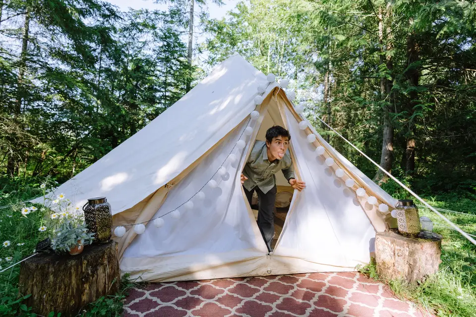 Young man sticking out his tongue and making a funny face while peeking out of a white canvas camping tent in a forested area with trees and greenery, decorated with string lights and lamps on tree stumps.
