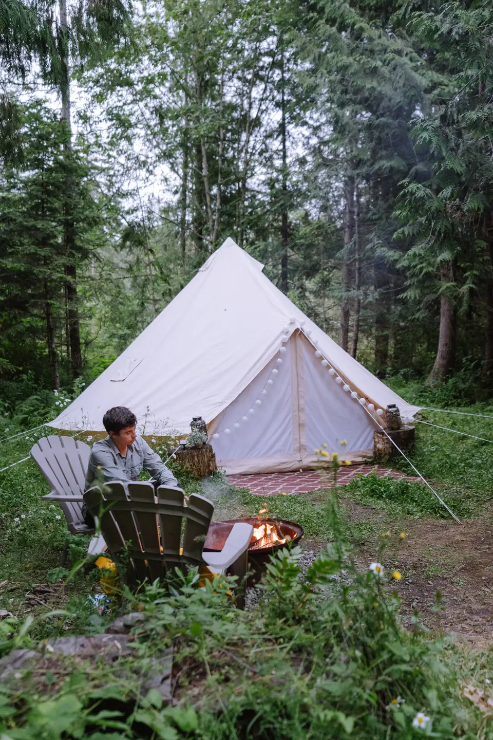 A person sitting in an outdoor chair near a campfire with a white canvas bell tent in the background, set in a forested area with surrounding greenery and trees.