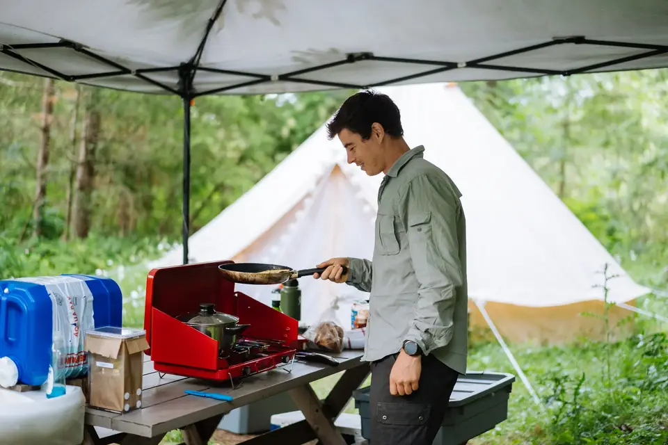 A man cooking on a portable stove in an outdoor camping area with a white tent in the background.