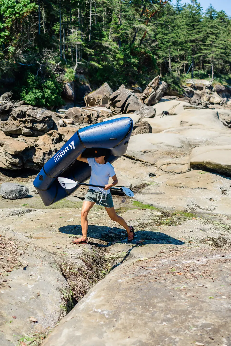 A person carrying a blue inflatable kayak and a paddle on a rocky riverbank with trees in the background.