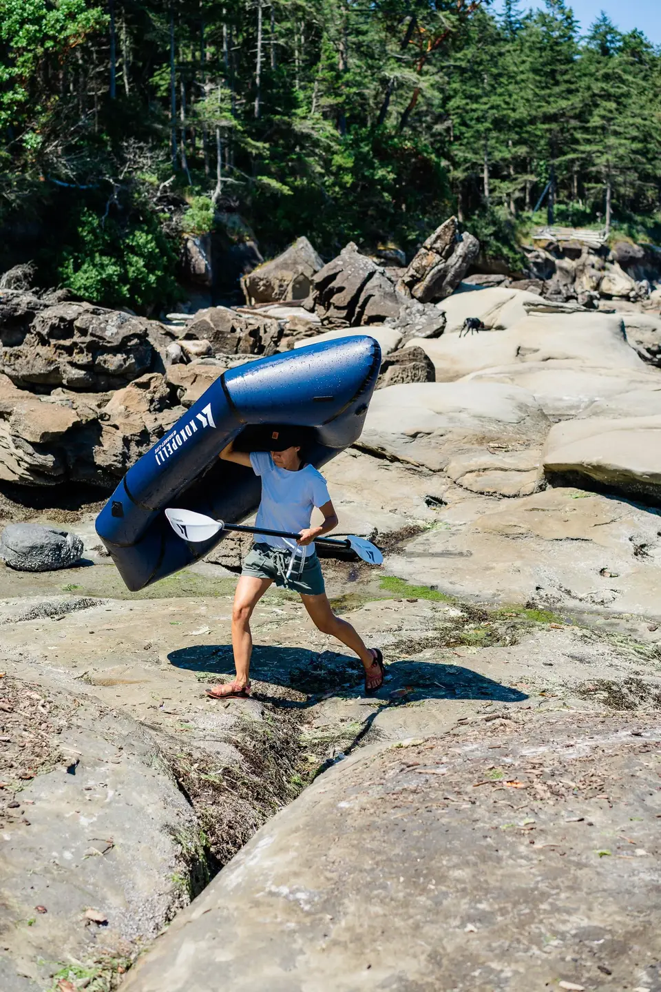 Carrying an inflated blue packraft across sun-bleached rocks on Hornby Island, British Columbia