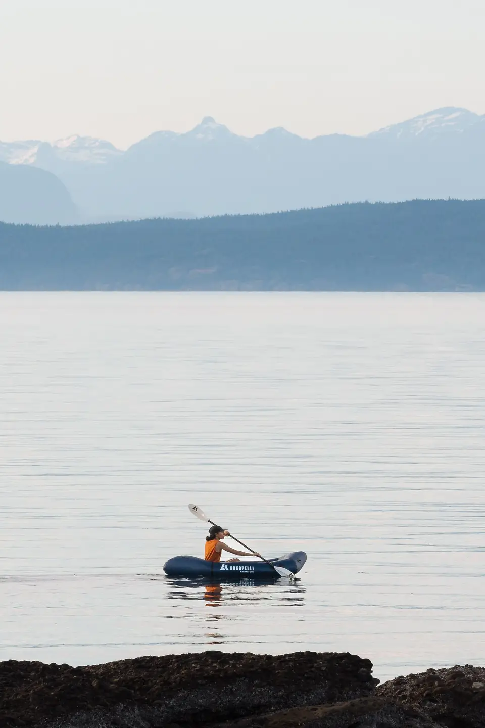 A person kayaking on calm water with mountains in the background.