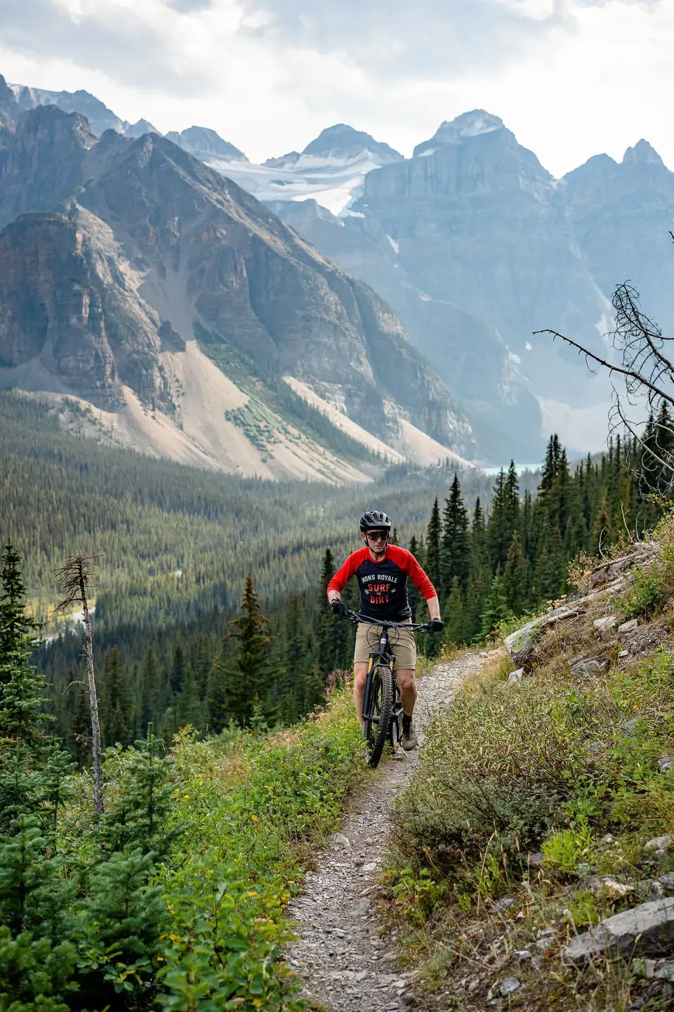 A rider coming down a narrow singletrack toward the camera below rocky peaks near Banff, Alberta