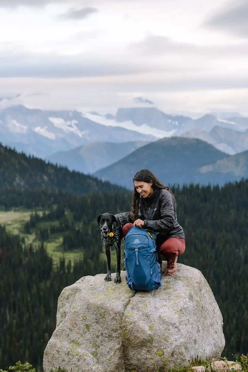 A hiker crouched on a boulder with Penelope and a blue pack, snowy ranges behind, in Revelstoke, British Columbia