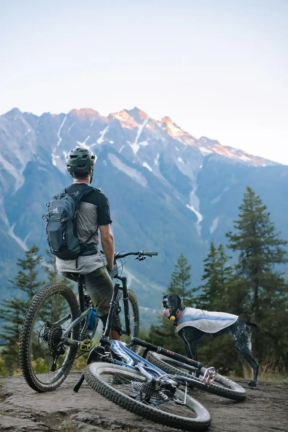 A rider with two bikes and Penelope on a rocky overlook facing jagged peaks in Whistler, British Columbia