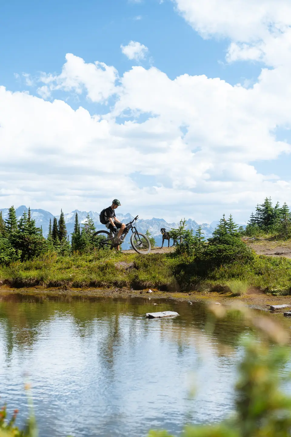 A mountain biker riding the far bank of a small alpine pond under summer clouds in Revelstoke, British Columbia