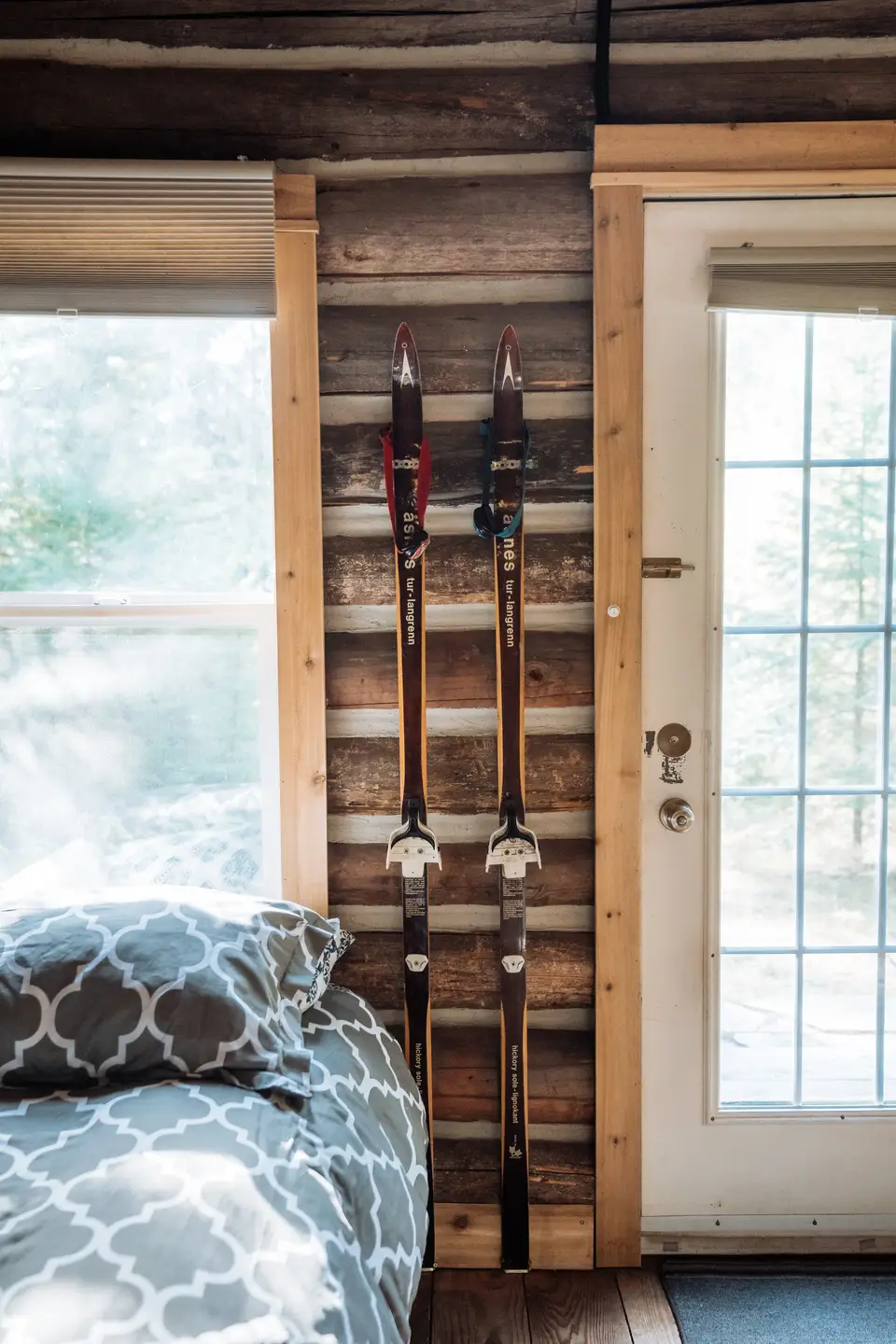 Two pairs of cross-country skis mounted on a wooden wall inside a room, next to a window and a door, with part of a bed with patterned bedding visible in the foreground.