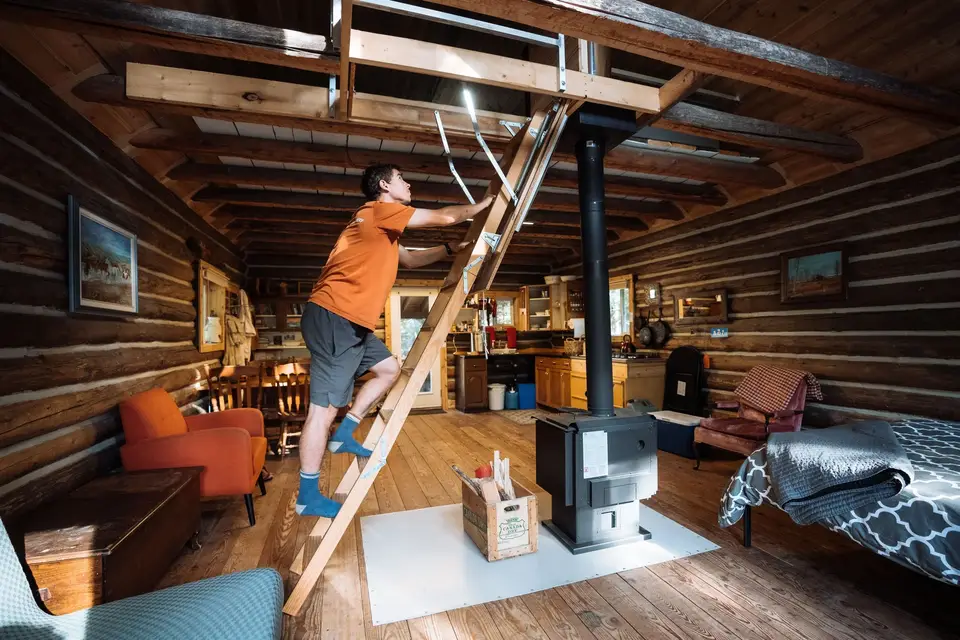 A man climbing a wooden ladder in a rustic log cabin, reaching towards the upper storage area of the ceiling, with a wood stove and cozy furniture visible in the room.