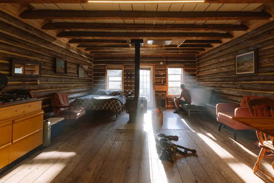 A man sitting on a bench near a window in a rustic log cabin interior with sunlight streaming in and a dog lying on the wooden floor.
