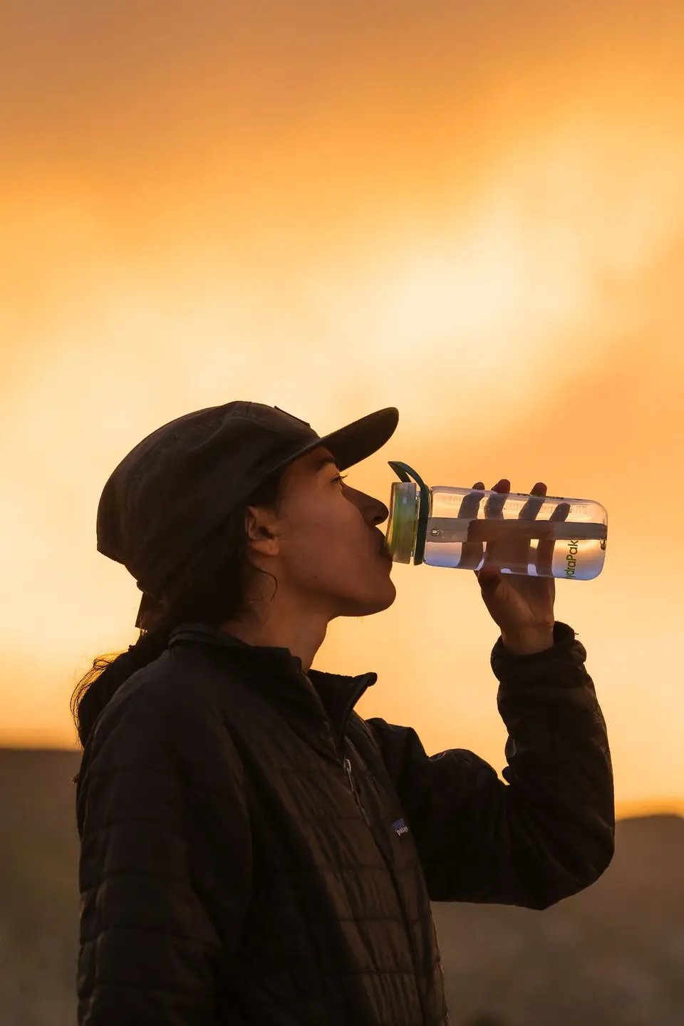 A woman wearing a dark jacket and cap drinks water from a clear bottle during sunset or sunrise.