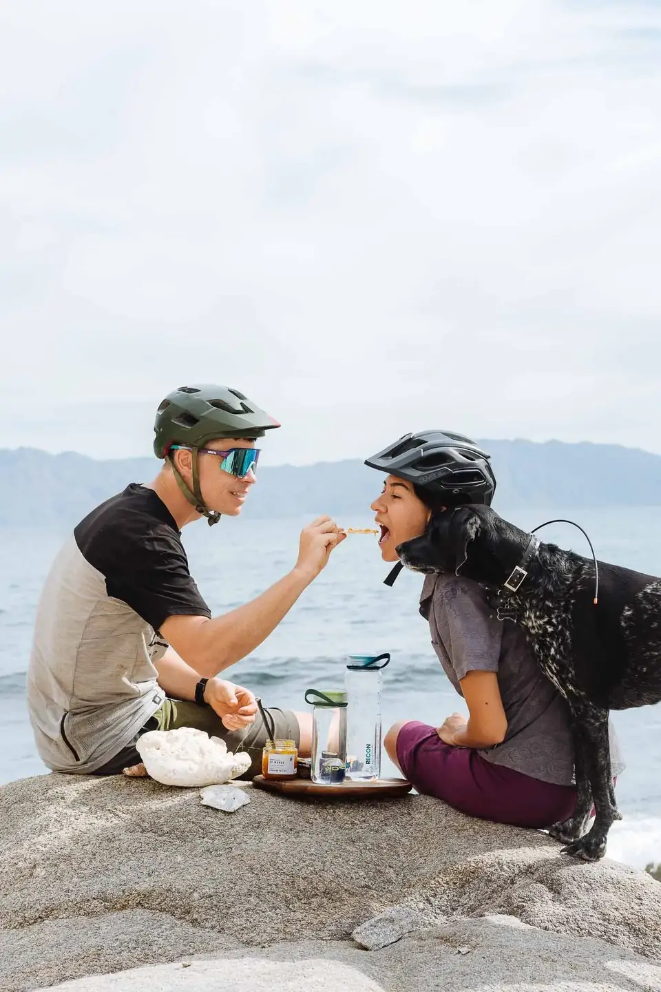 A man feeding a woman a snack while both wear helmets, sitting on rocks by the water with mountains in the background and a dog with a harness nearby.