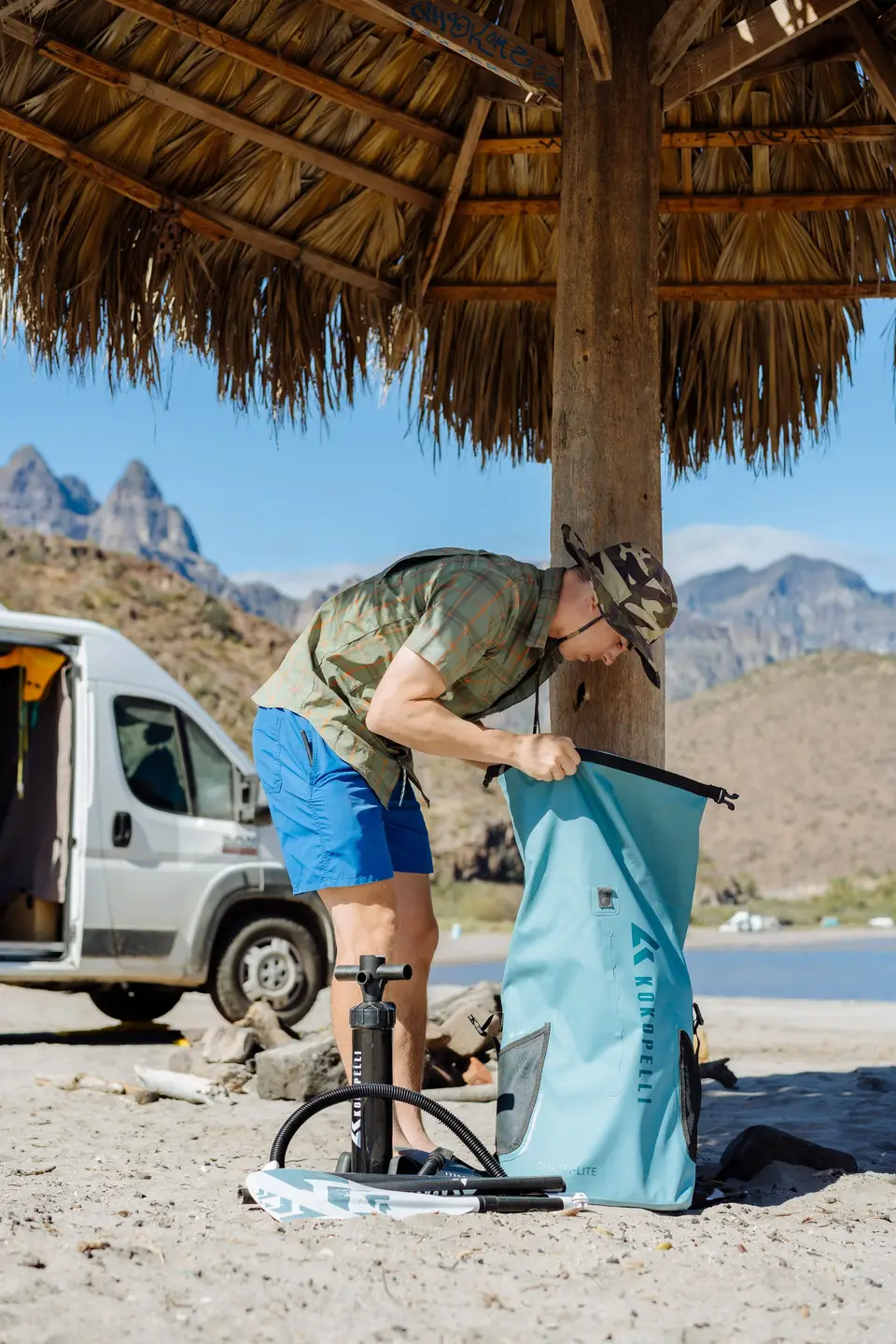 A man prepares a backpack and stands under a tiki hut on a sandy beach with mountains in the background.