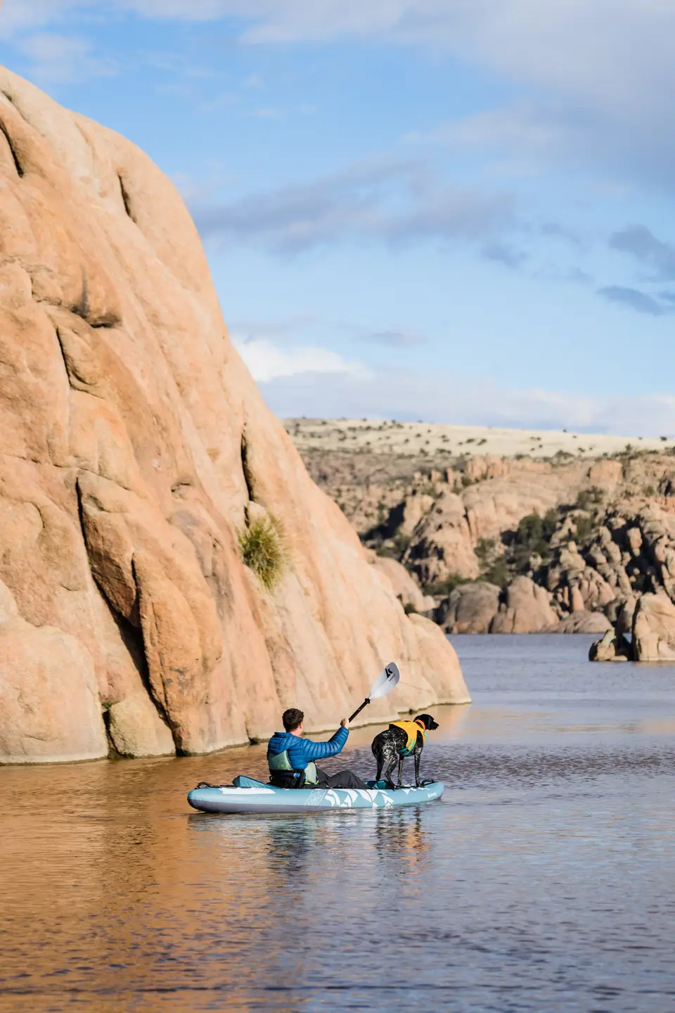Man kayaking with his dog on a river in a rocky canyon under a partly cloudy sky.
