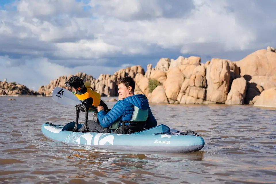 A man and a dog on a paddleboard in a water body with rocky formations and a cloudy sky in the background.