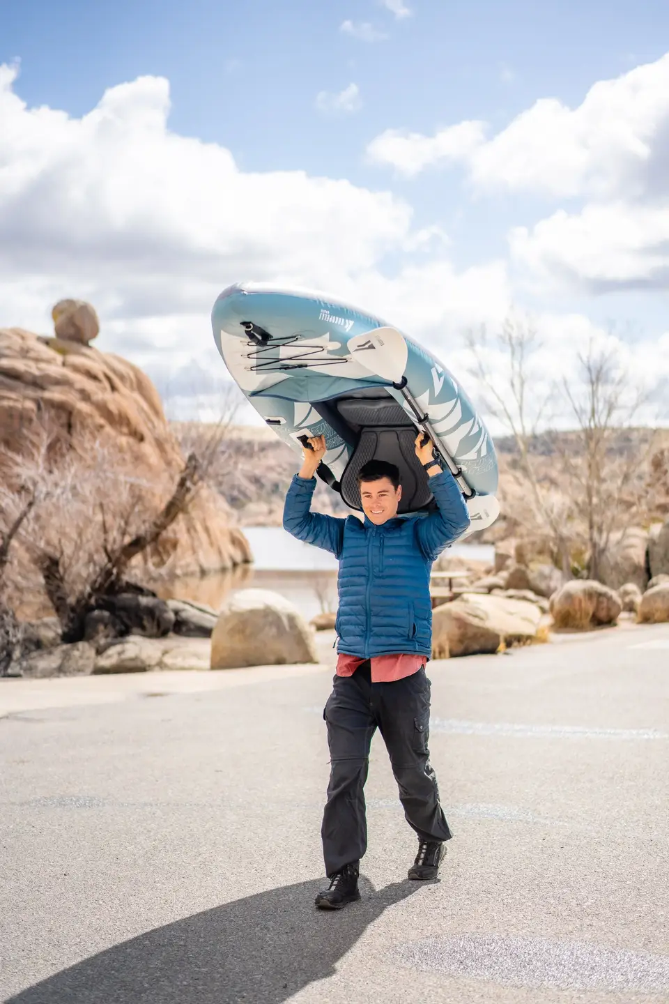 A smiling man carrying a kayak on his shoulders outdoors on a paved area with rocks and leafless trees, under a partly cloudy sky.