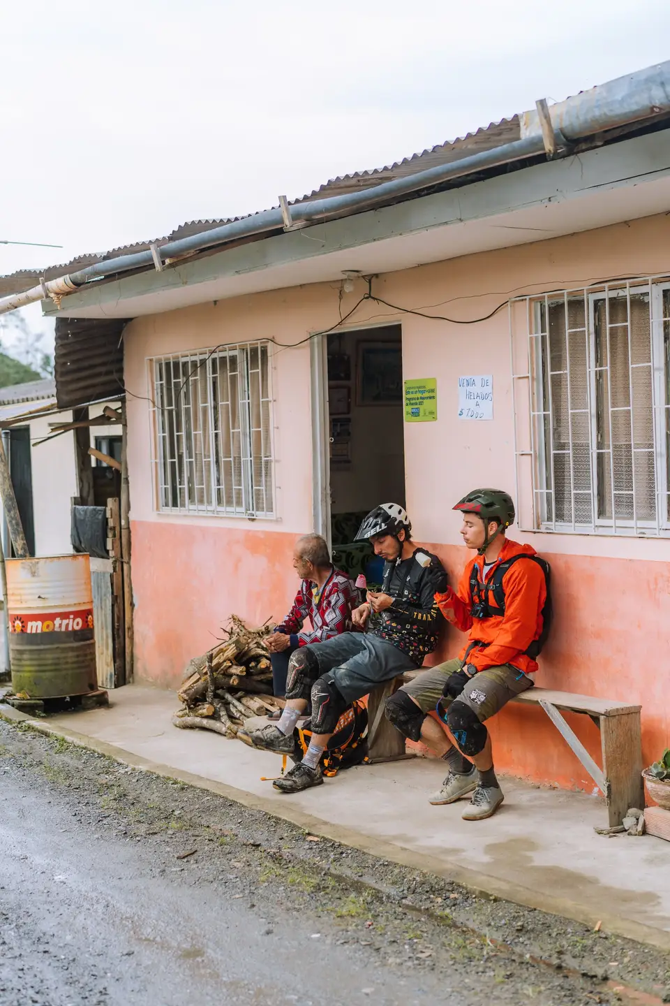 Two mountain bikers and a local man on a bench outside a peach storefront in rural Colombia