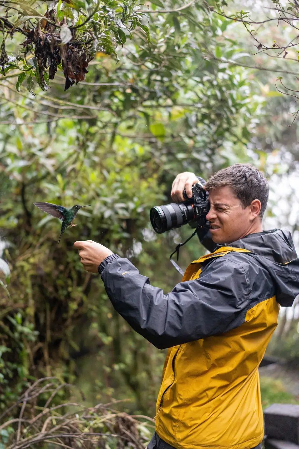 Evan in a rain jacket raising a camera in leafy woods in Colombia