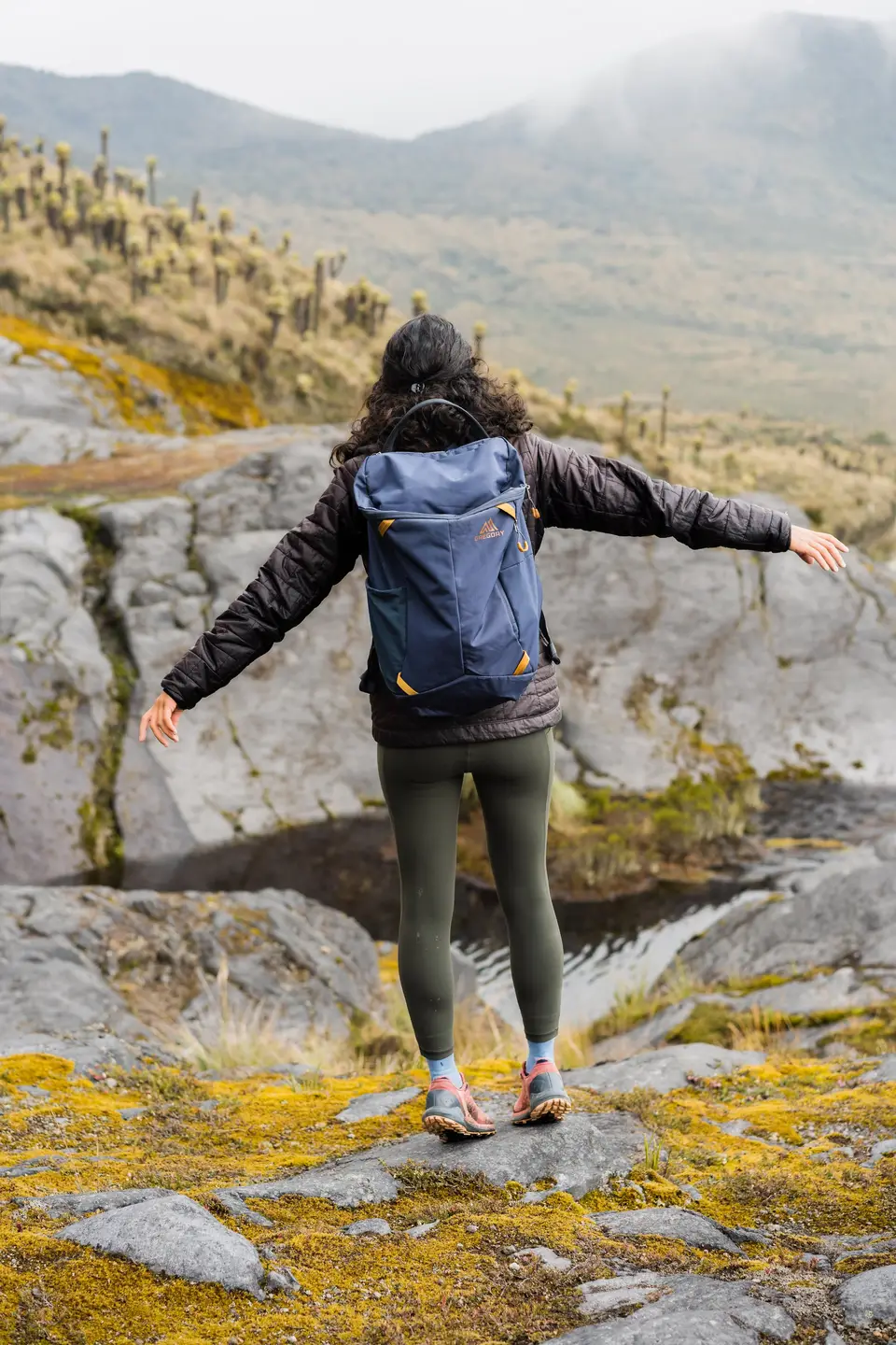 Melanny on a mossy ledge in the Colombian páramo, slate-blue Gregory pack on her back, arms open