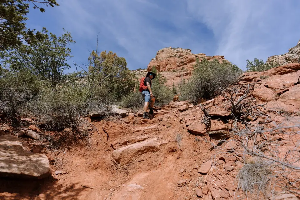 A hiker climbing the steep red dirt trail toward the Keyhole Cave