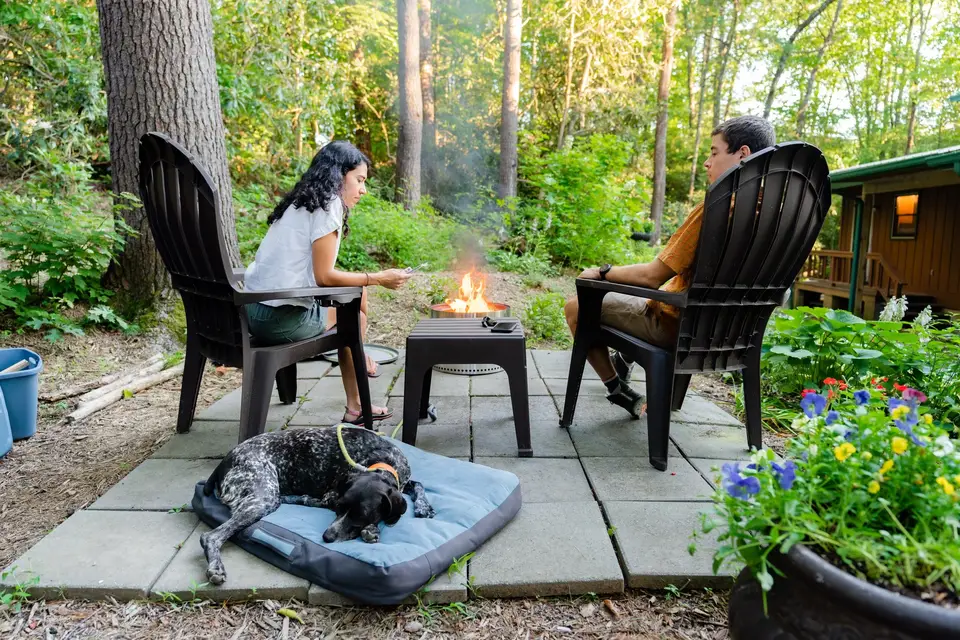 Two people sitting outdoor around a small fire pit in a wooded backyard, with a dog lying on a bed nearby. One person is checking their phone while the other is relaxing with arms crossed.