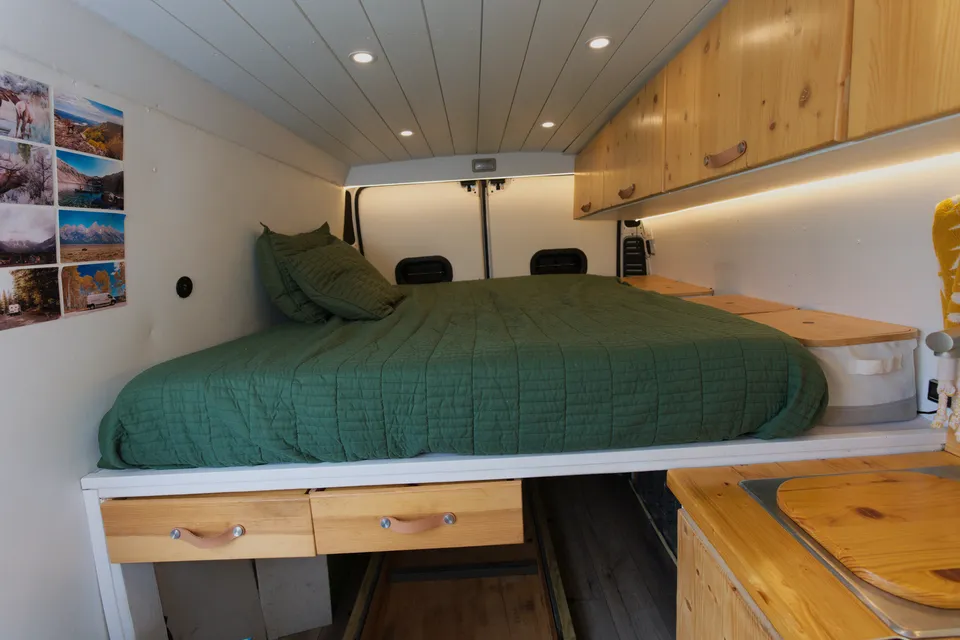 Raised bed with a green comforter under wood cabinets and LED strip lighting inside the van